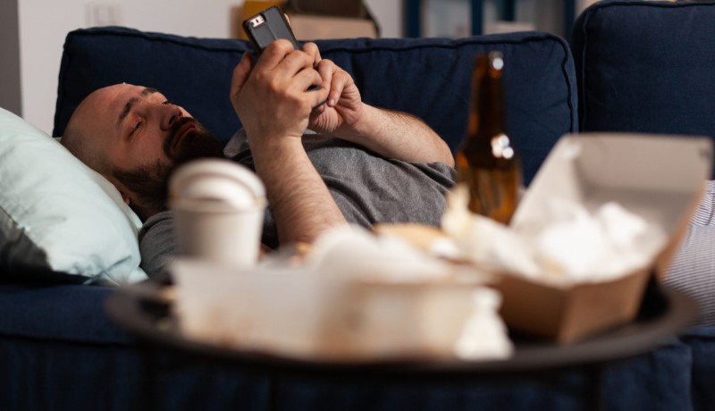 A bearded man lounges on a blue couch, looking at his phone