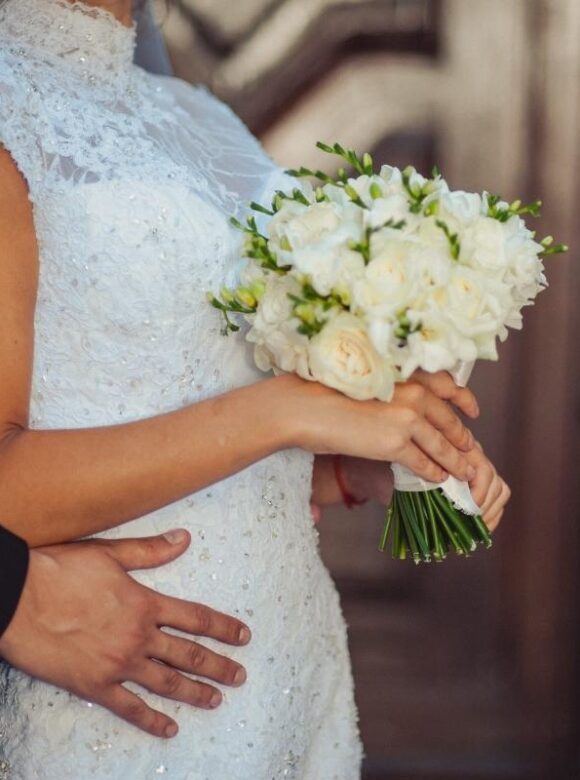 Bride wearing a white lace wedding dress and holding a bouquet of white roses while her partner embraces her from behind.
