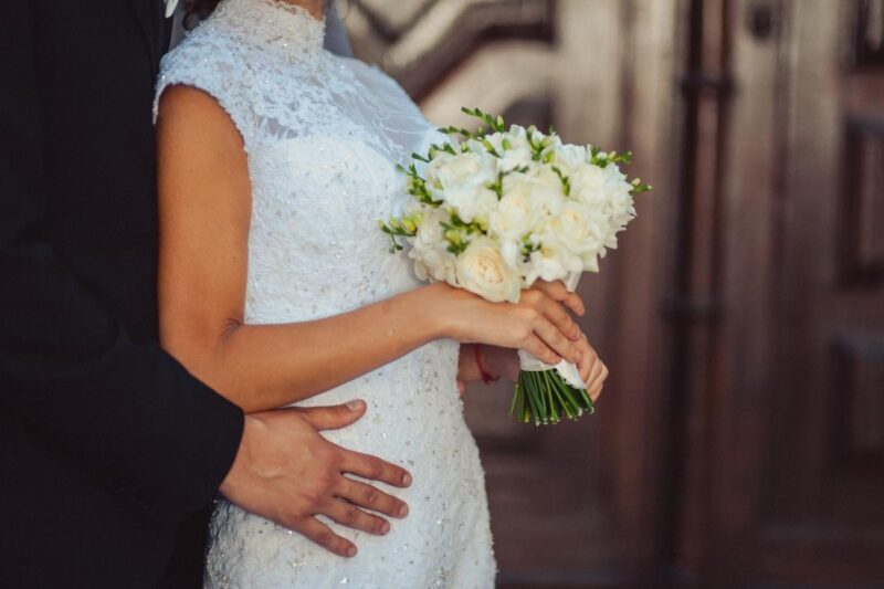 Bride wearing a white lace wedding dress and holding a bouquet of white roses while her partner embraces her from behind.