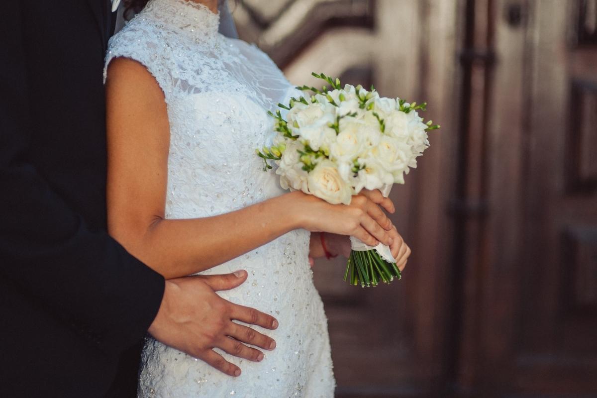 Bride wearing a white lace wedding dress and holding a bouquet of white roses while her partner embraces her from behind.