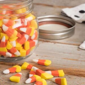 Candy corn fills a glass jar with several pieces scattered on the wooden table.