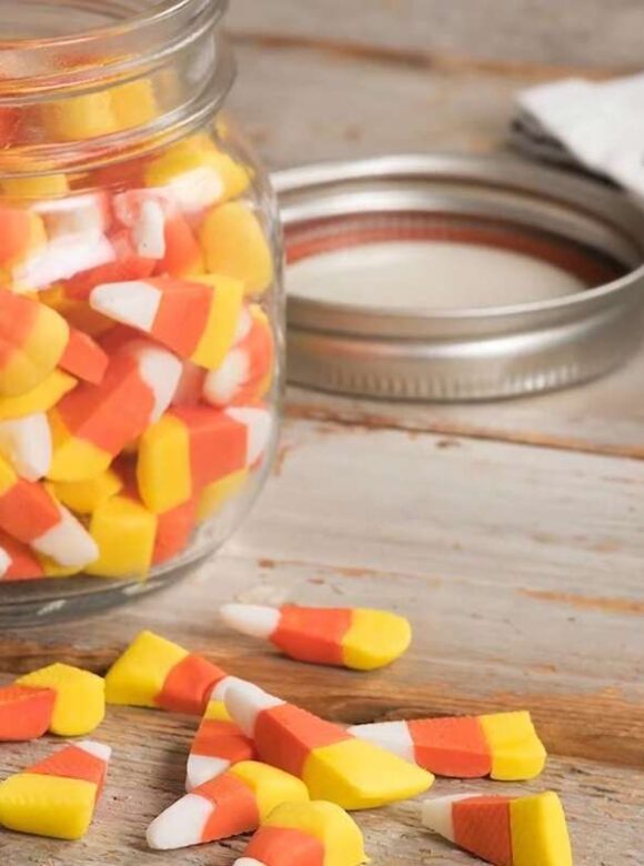 Candy corn fills a glass jar with several pieces scattered on the wooden table.
