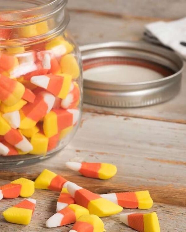 Candy corn fills a glass jar with several pieces scattered on the wooden table.