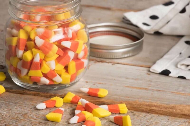Candy corn fills a glass jar with several pieces scattered on the wooden table.