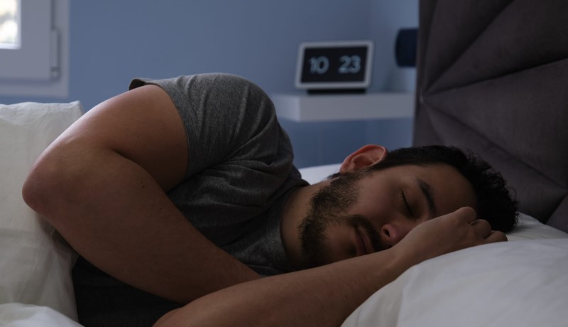 A bearded man sleeps curled on his side beneath white bedding in a dim bedroom