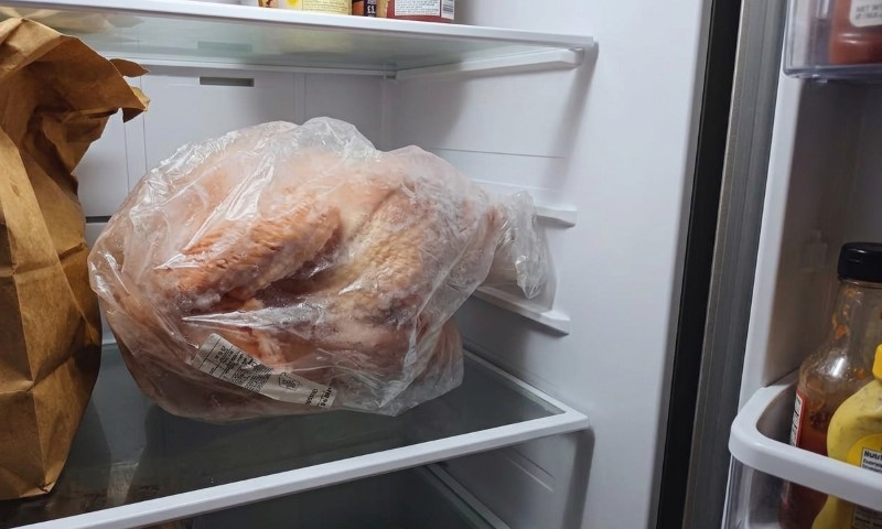 A plastic-wrapped roasted chicken sits on a refrigerator shelf beside a brown paper bag