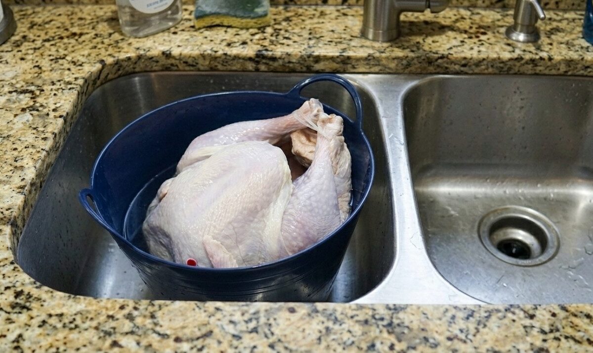A raw whole turkey sits breast-up in a dark blue roasting pan inside a stainless-steel kitchen sink