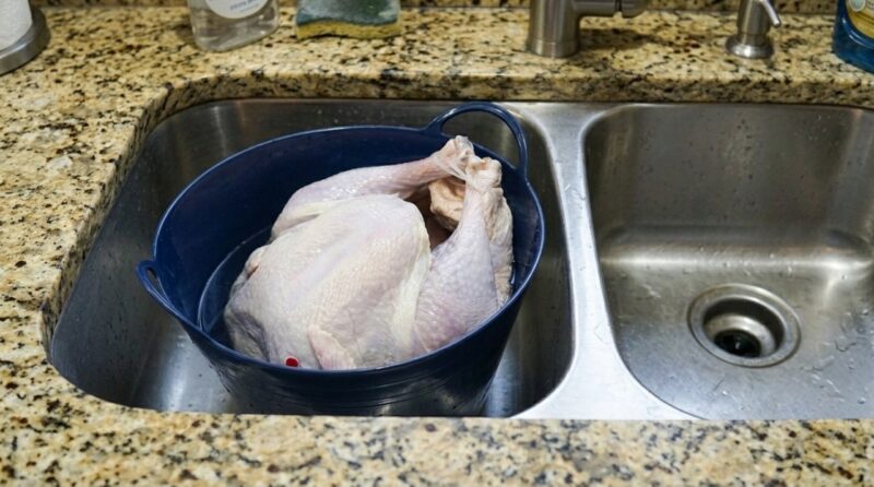 A raw whole turkey sits breast-up in a dark blue roasting pan inside a stainless-steel kitchen sink
