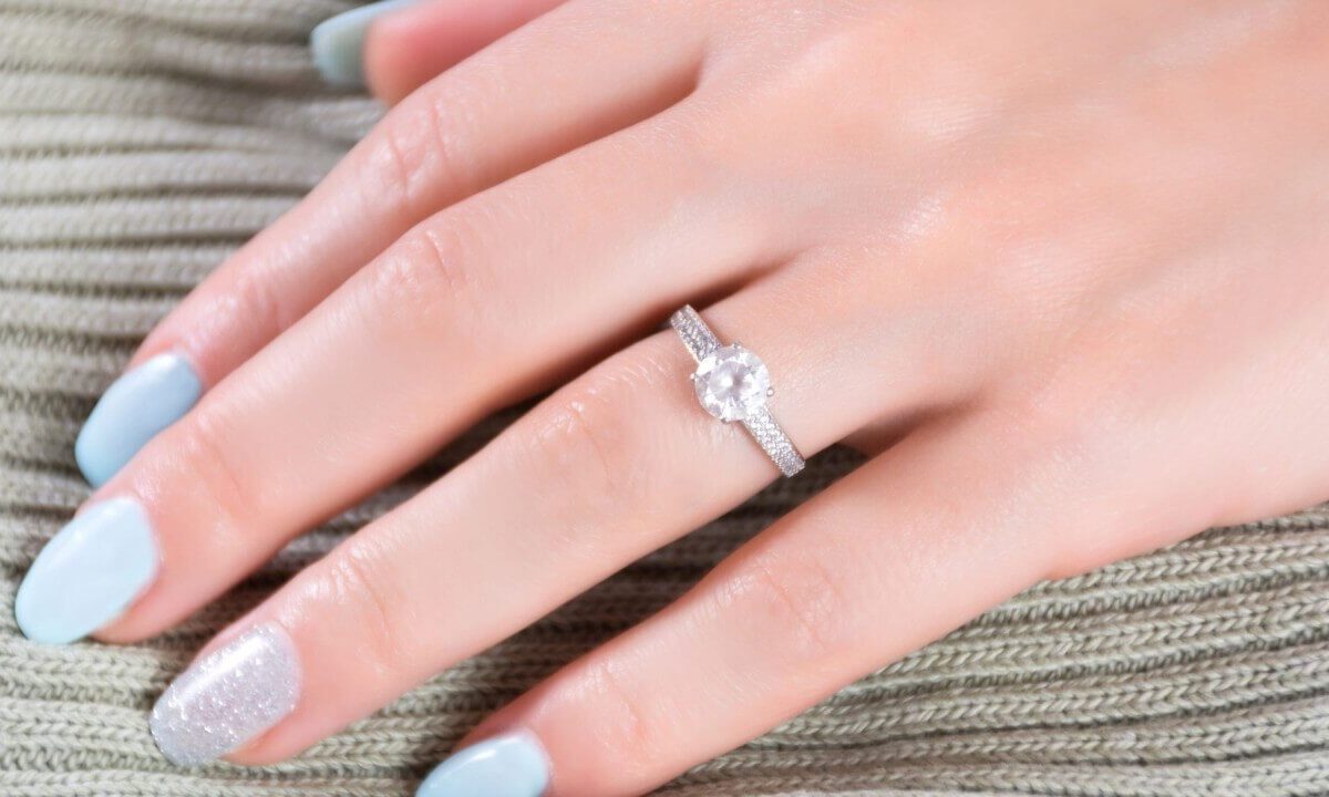 A hand with pale blue and glitter-polished nails wears a sparkling diamond engagement ring