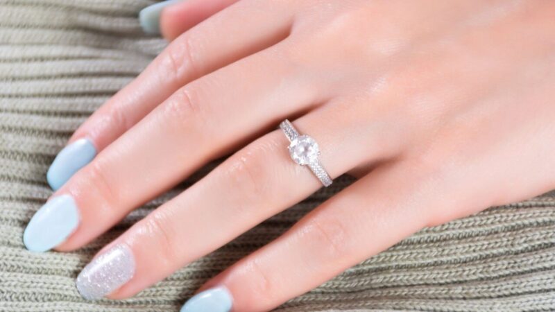 A hand with pale blue and glitter-polished nails wears a sparkling diamond engagement ring