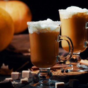 Two pumpkin spice lattes topped with whipped cream sit beside pumpkins and cinnamon sticks.