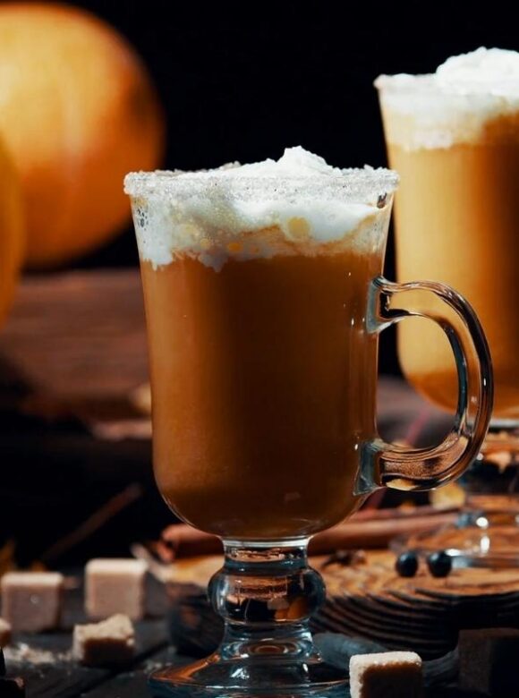 Two pumpkin spice lattes topped with whipped cream sit beside pumpkins and cinnamon sticks.