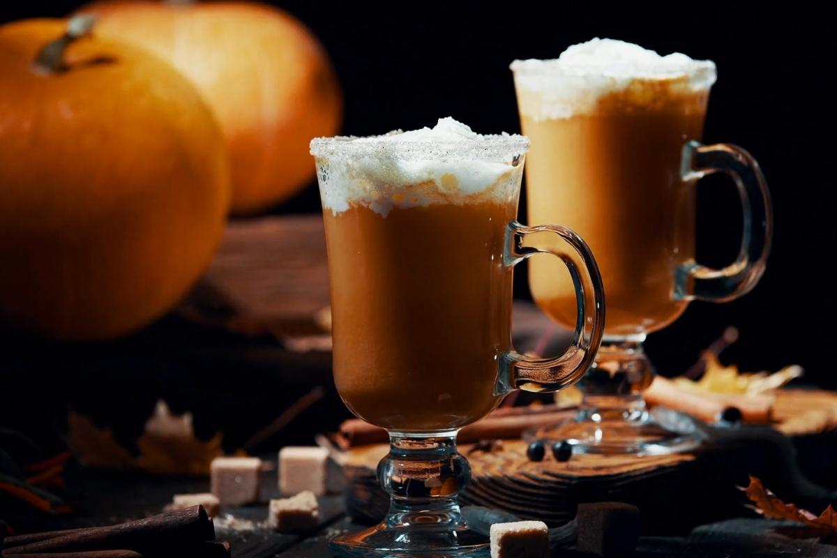 Two pumpkin spice lattes topped with whipped cream sit beside pumpkins and cinnamon sticks.