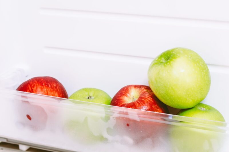 A clear refrigerator drawer holds red and green apples, including a bright green apple in front
