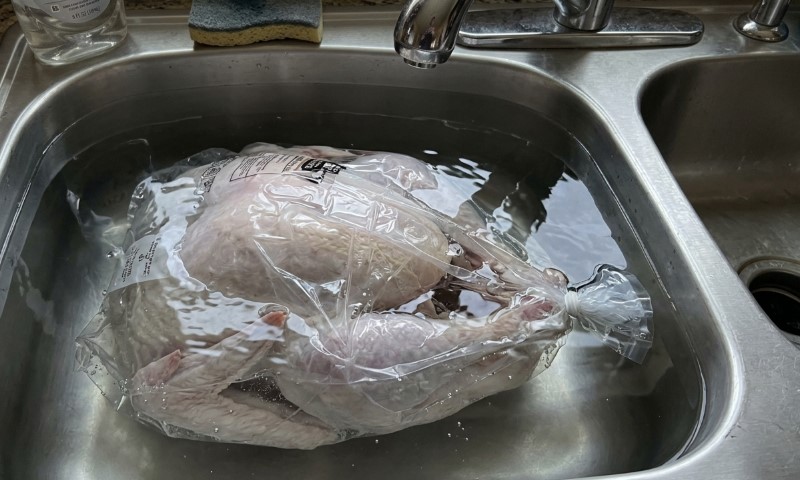 A raw whole turkey sealed in plastic thaws in a water-filled stainless steel kitchen sink