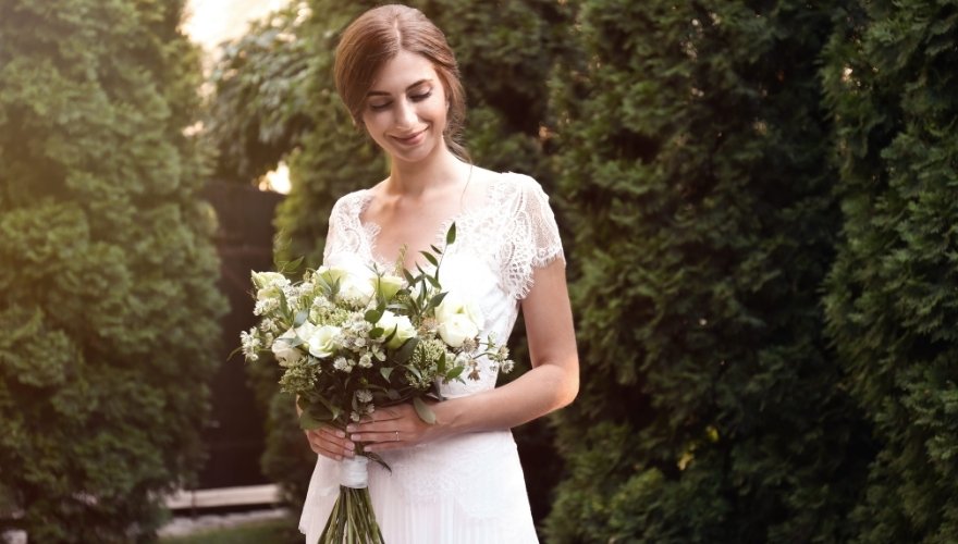 Bride wearing a white lace wedding dress and holding a bouquet of white roses and greenery in an outdoor garden.