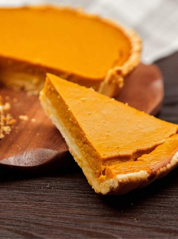 Homemade pumpkin pie with a slice cut and placed beside it on a wooden table, illustrating a dessert that requires refrigeration for safe storage.
