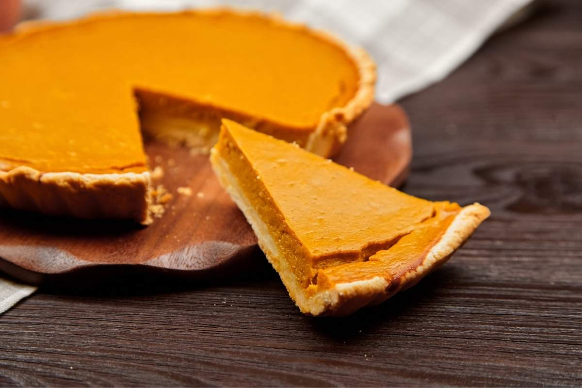 Homemade pumpkin pie with a slice cut and placed beside it on a wooden table, illustrating a dessert that requires refrigeration for safe storage.